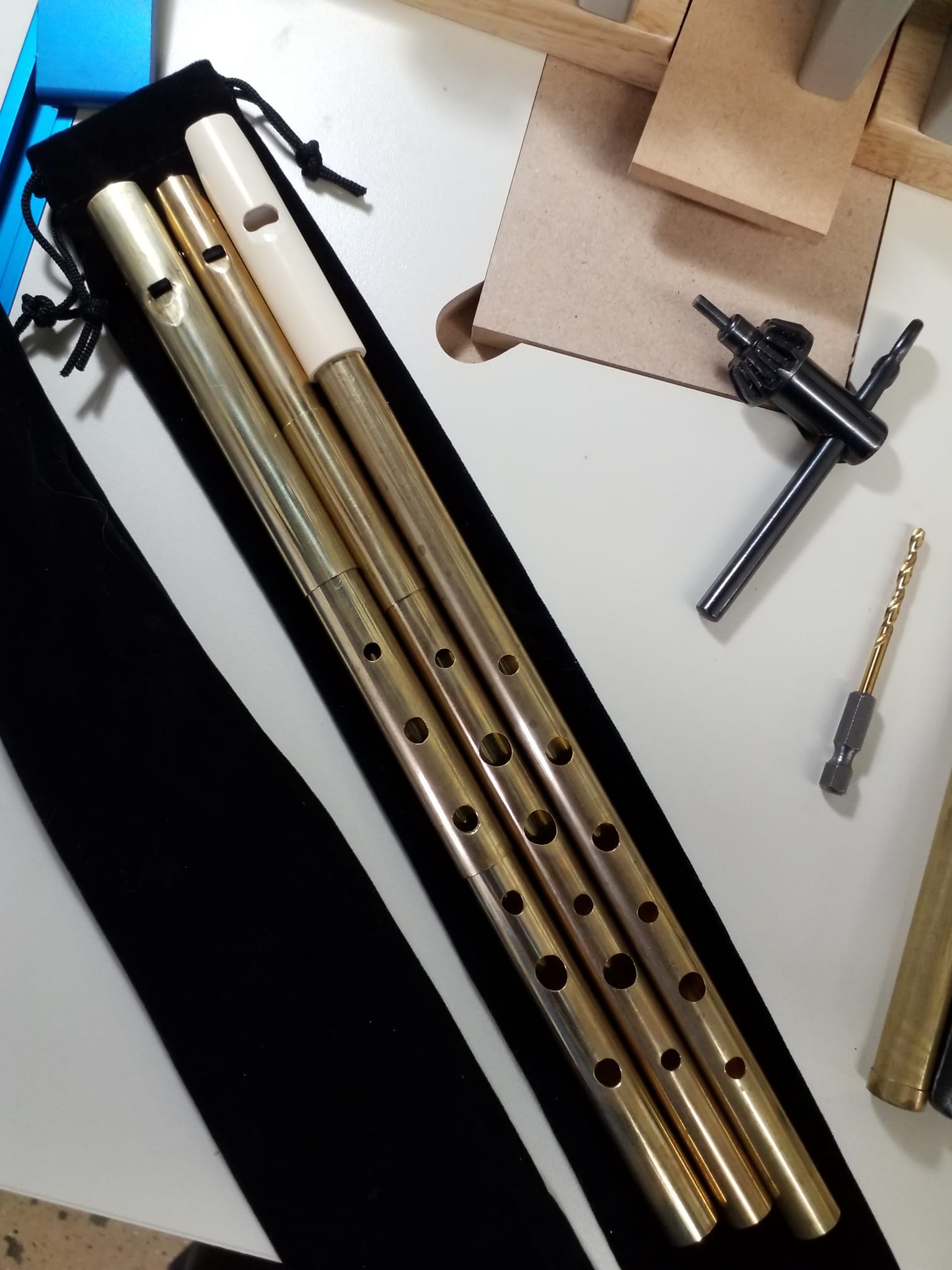 Three MackBeth brass whistles resting on a black velvet case.  One whistle has a white headpiece.  The case is on a white drill press table.  A drill bit and a drill chuck key are visible to the right.